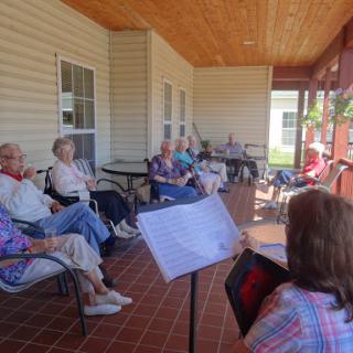 A group of seniors enjoying a musical performance on a sunny porch.