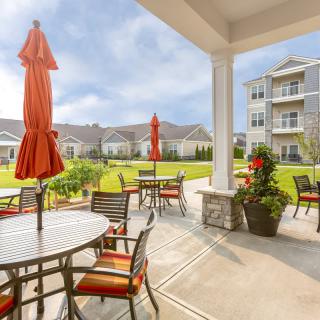 A well-maintained outdoor patio area with tables, chairs, and red umbrellas overlooking landscaped grounds and nearby apartment buildings.