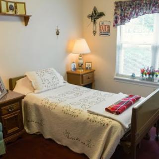 A cozy and simple bedroom featuring a single bed with a decorative blanket, a nightstand with a lamp, and warm natural light coming through the window.