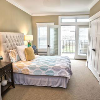 A cozy bedroom featuring a neatly made bed with colorful pillows, a bedside table with a lamp, and large windows letting in natural light.
