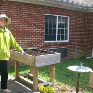 A person in a sun hat stands next to a raised garden bed outside a brick building.