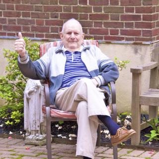 An elderly man sitting comfortably in a garden chair, giving a thumbs up gesture.