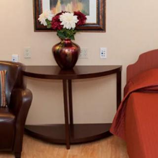 A cozy bedroom corner featuring a leather chair, a wooden half-moon table with a decorative vase of flowers, and a neatly made bed with an orange bedspread.