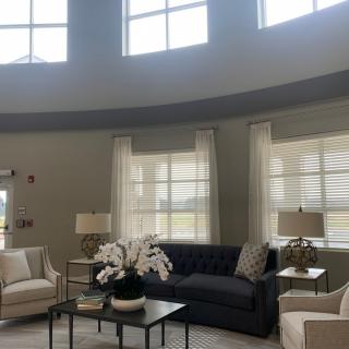 A spacious and elegantly designed living room with large windows, featuring a dark sofa, light-colored chairs, lamps, and a central table adorned with flowers.