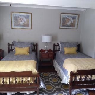 A cozy bedroom with twin beds featuring yellow and gray bedding, a central nightstand with a lamp, and decorative wall art above.