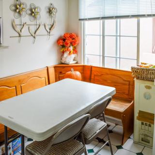 A cozy kitchen nook with a white table, metal chairs, and wooden bench seating by a window adorned with decorative flowers.