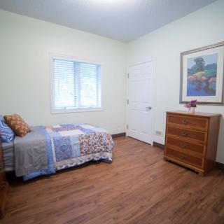 A minimalist bedroom features a single bed, wooden floors, and a dresser beneath a framed painting.