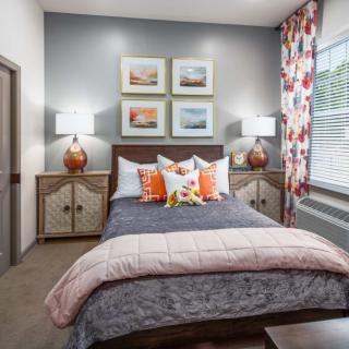 A stylish bedroom featuring a well-made bed with decorative pillows, flanked by bedside tables with lamps. The walls display framed artwork, and a large window allows natural light to complement the floral curtains.