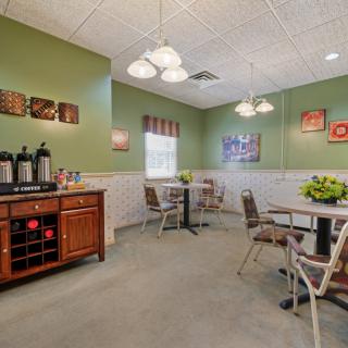 A cozy dining area with a coffee station, featuring tables and chairs under warm lighting, complemented by green walls and decorative art.