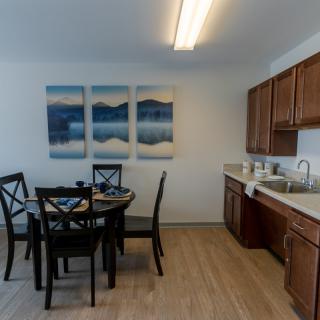 A modern dining area with a table and chairs, adjacent to a kitchen with wooden cabinets and a sink, featuring a triptych artwork on the wall.