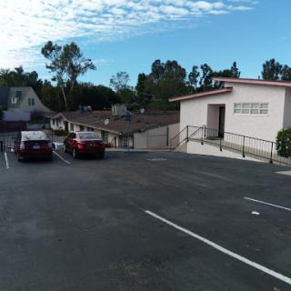 A view of an empty parking lot with a few parked cars and surrounding greenery under a partly cloudy sky.