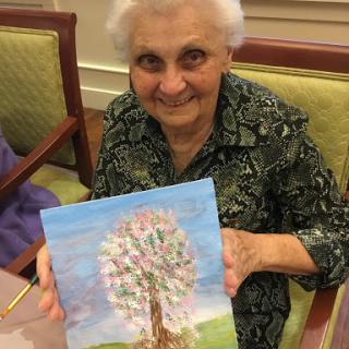 An elderly woman proudly displays her painting of a tree, showcasing her artistic talent and joy.