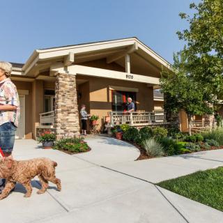 A person walking a dog in front of a house with people socializing on the porch.