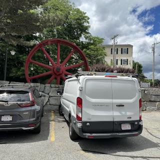 A parked white van in front of a large red historic wheel, with a backdrop of a cloudy sky and a building.
