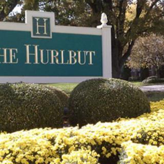 A green and white sign for 'The Hurlbut' surrounded by neatly trimmed bushes and yellow flowers.