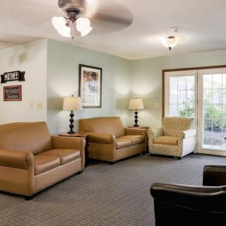 A cozy living room with a ceiling fan, sofas, chairs, lamps, and a view of the outdoors through large windows.