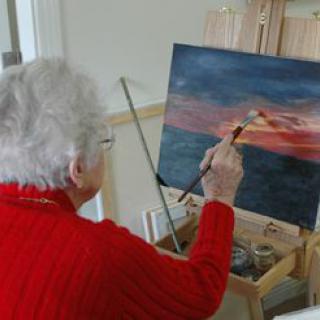 An elderly woman painting a landscape at an easel.