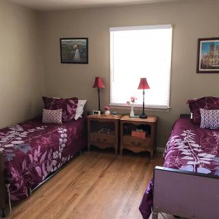 A cozy bedroom with two twin beds featuring purple floral bedding, a shared nightstand with red lamps, and framed artwork on the walls.