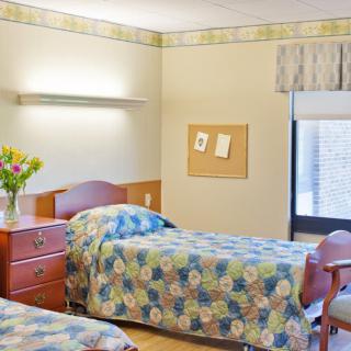 A hospital room with two twin beds, a wooden dresser with flowers, and a window with blinds.