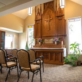 A serene chapel interior with chairs facing an altar adorned with a wooden cross, surrounded by plants.