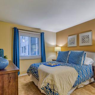A cozy bedroom with blue and beige decor, featuring a comfortable bed, a lamp, and beautiful artwork on the walls.