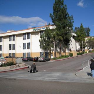 A view of a residential apartment building with people on the sidewalk and trees nearby.