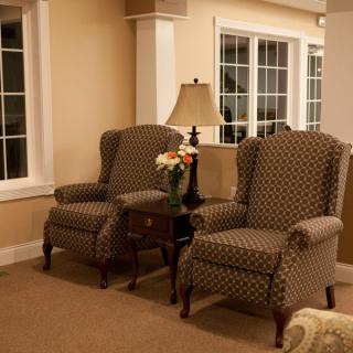 A cozy living room with two patterned armchairs and a small table between them, adorned with a lamp and flowers.