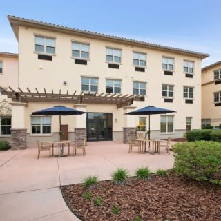 A modern three-story building with a patio area featuring outdoor seating and sunshades.