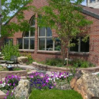 A brick building with large windows surrounded by a landscaped garden featuring colorful flowers and trees.