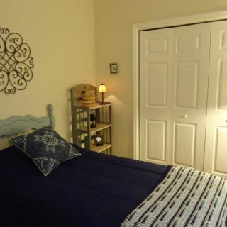 A cozy bedroom with a neatly made bed, decorated with pillows and a throw blanket. A small shelf with a lamp is placed beside the bed, and a closed closet is visible in the background.