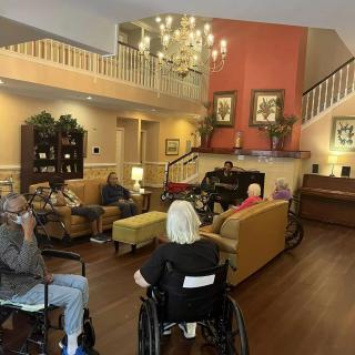A cozy nursing home lounge where residents are gathered, some in wheelchairs, enjoying a communal space with sofas and a chandelier.
