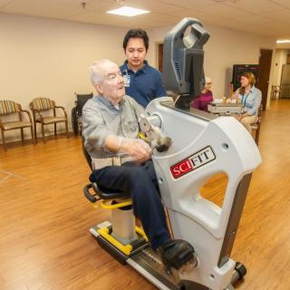 An elderly person exercises on a stationary machine under the guidance of a healthcare professional.