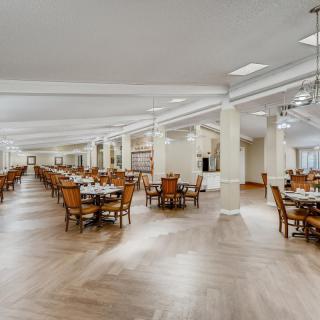 A spacious dining hall with wooden flooring, filled with neatly arranged tables and chairs.