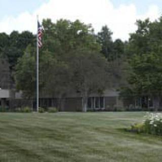 A building with an American flag on a lush green lawn, surrounded by trees under a clear sky.