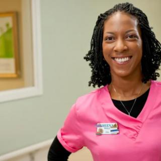 A nurse smiling in a hospital setting, wearing pink scrubs and a name badge.