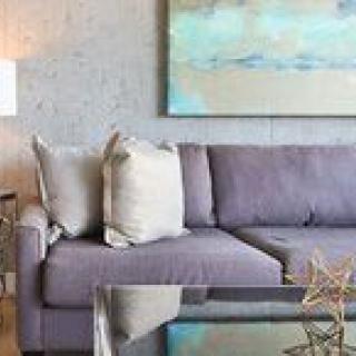 A modern living room features a gray sofa with cushions, an abstract painting on the wall, and a glass coffee table.