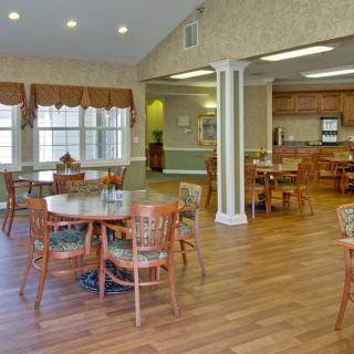 A spacious dining area with wooden tables and chairs, adjacent to a kitchen, featuring natural light and a wooden floor.