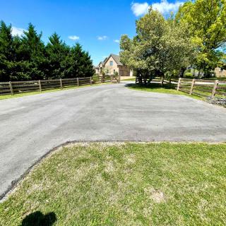 A paved driveway leading to a house, surrounded by trees and a wooden fence under a clear blue sky.