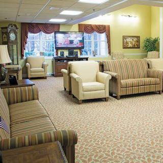 A cozy living room with multiple sofas and chairs arranged around a television, featuring carpeted flooring and large windows.