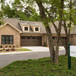 A brick residential house with a driveway and garage, surrounded by trees, featuring a real estate sign in the yard.