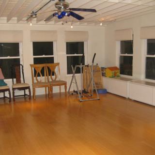 An empty room with hardwood floors, featuring a few chairs and exercise equipment near large windows.