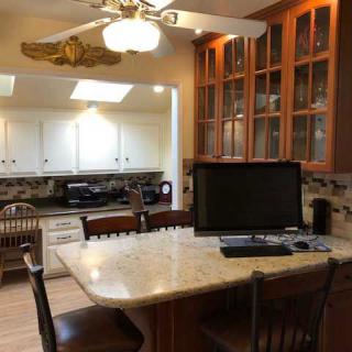 A kitchen with a central island featuring a computer monitor and seating, surrounded by wooden cabinets and a ceiling fan.