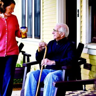 An elderly man sitting on a porch, holding a walking cane, while conversing with a woman standing nearby.