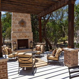 An outdoor patio with comfortable seating around a fireplace under a pergola, surrounded by trees.