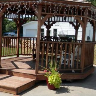 A wooden gazebo decorated with fairy lights and surrounded by a potted plant on a sunny day.
