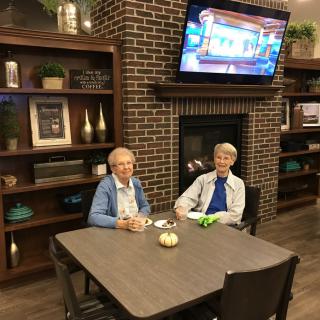 Two elderly individuals sitting at a table in a cozy room with a TV and fireplace.