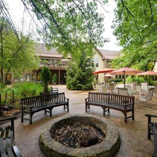 A serene courtyard featuring wooden benches surrounding a stone fire pit, with lush greenery and outdoor seating areas.