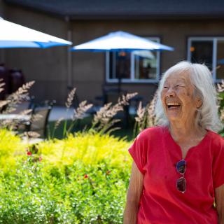 An elderly woman enjoys a sunny day in a garden, smiling brightly.