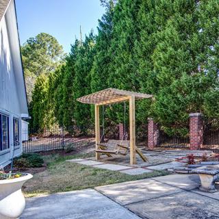 A backyard view featuring a small wooden pergola, surrounded by lush green trees and a paved patio area.