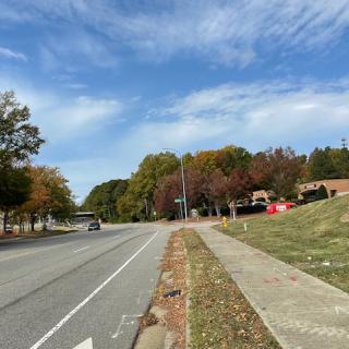 A scenic road bordered by a sidewalk, autumn trees, and nearby buildings under a partly cloudy sky.